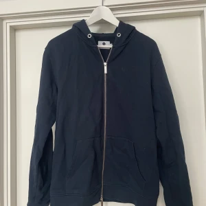 Marinblå hoodie från NN07 i nyskick - Säljer en stilren svart hoodie från NN07 i storlek S. Den har en praktisk dragkedja framtill och en klassisk huva. Perfekt för en avslappnad look. Använd ett fåtal gånger därmed i princip i nyskick. Nypris ca 1500kr