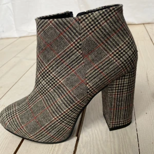 Rutig stövlett med klack - Kaxiga rutiga stövletter från Ellos med 10 cm hög klack och dragkedja på sidan. Skorna har ett klassiskt rutmönster i grått, svart och rött, vilket ger dem en stilren look. Fodret har en skada på ett ställe, se bild. Storlek 38 men han nog funka bra för en 37a också. Använda ett fåtal gånger. 