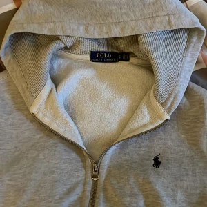 Grå hoodie från Polo Ralph Lauren - Säljer en stilren grå hoodie från Polo Ralph Lauren med dragkedja och broderad logga på bröstet. Tröjan har en mjuk insida och praktiska fickor framtill. Perfekt för en avslappnad look. Inga defekter, priset kan diskuteras vid snabb affär💯