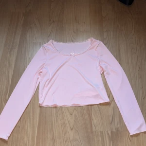 Rosa långärmad topp med spetsdetaljer - Söt rosa långärmad topp med spetsdetaljer runt halsringningen och en liten rosett framtill. Perfekt för en romantisk look. Toppen är croppad och har en mjuk och bekväm passform.