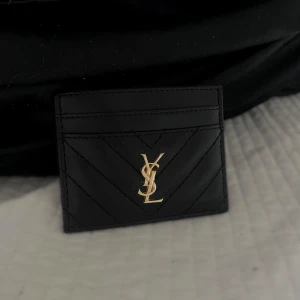 Svart kortfodral från YSL - Snyggt och stilrent svart kortfodral från YSL med guldlogga framtill. Perfekt för att hålla dina kort organiserade med stil. Den quiltade designen ger en lyxig känsla. 