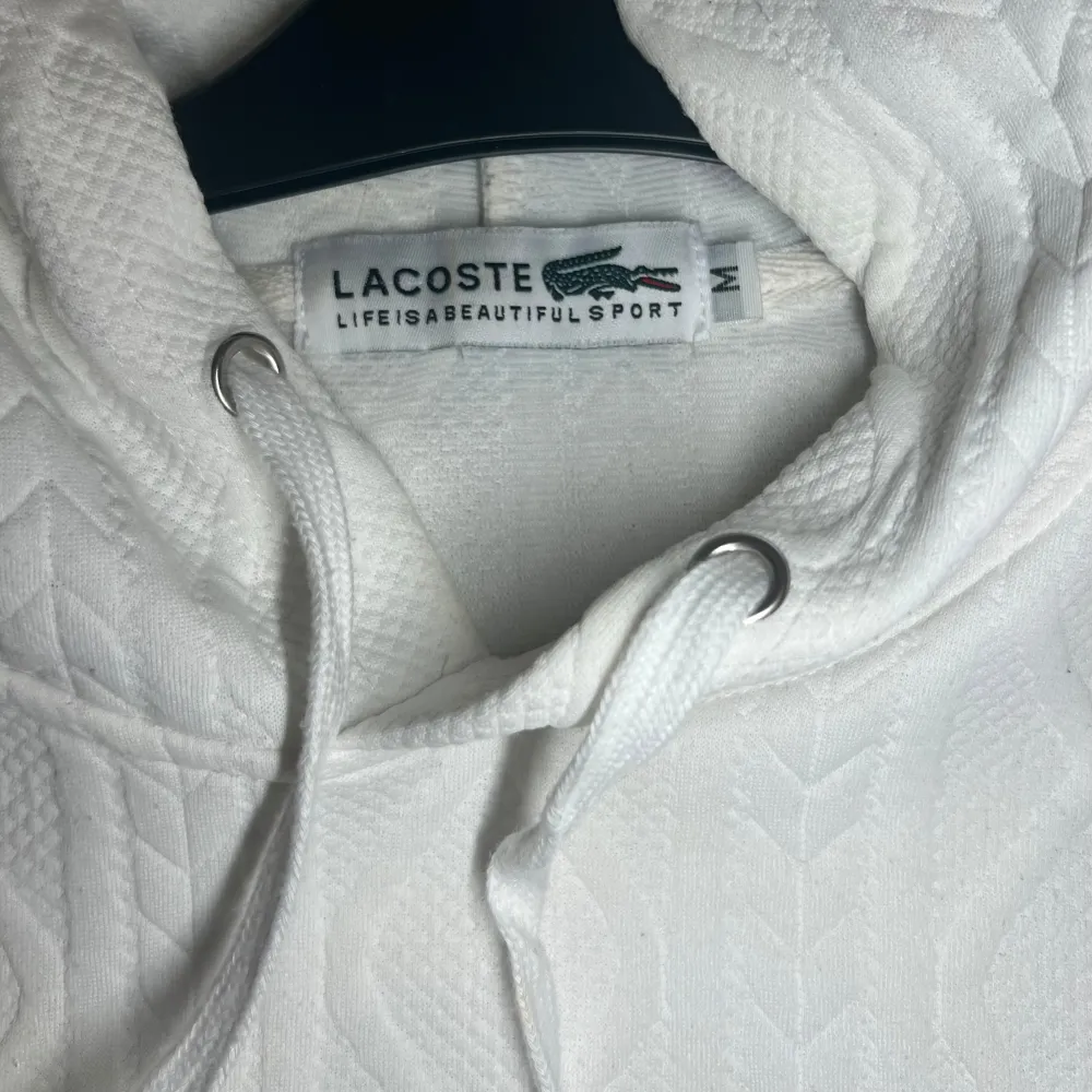 Säljer en stilren vit hoodie från Lacoste med det klassiska krokodilmärket på bröstet. Hoodien har ett snyggt mönster. Perfekt för en avslappnad och slim look. . Neuletakit & Villapaidat.