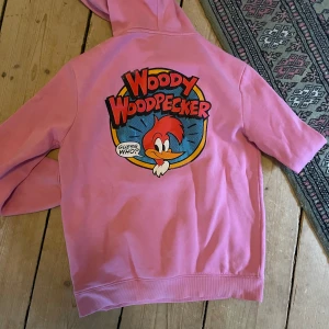 Rosa Woody Woodpecker hoodie från H&M - Säljer en rosa hoodie från H&M med ett stort Woody Woodpecker-tryck på ryggen och ett mindre tryck på bröstet. Hoodien har en klassisk design med huva och ribbade muddar vid ärmslut och nederkant. Perfekt för dig som gillar retrostil! 💖
