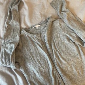 Grå kofta från Gina Tricot - Säljer en stilren grå kofta från Gina Tricot. Koftan har långa ärmar och knappar framtill, perfekt för lager på lager-stil. Den är mjuk och bekväm, idealisk för kyligare dagar. Passar både till jeans och kjol för en avslappnad look.