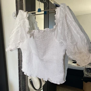 Vit blus med puffärmar - Säljer en söt vit blus med puffiga kortärmar och elastisk struktur. Blusen har en fyrkantig halsringning och volangdetaljer vid ärmslut och nederkant. Perfekt för en somrig look!