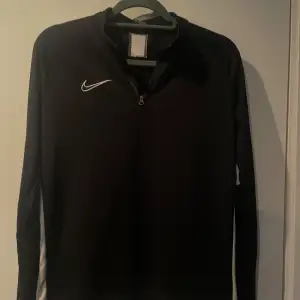 Säljer en svart långärmad tröja från Nike med dragkedja vid halsen. Tröjan har en vit Nike-logga på bröstet och vita detaljer längs sidorna. Perfekt för träning eller vardagsbruk.