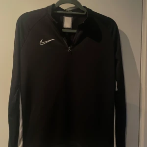 Svart långärmad tröja från Nike - Säljer en svart långärmad tröja från Nike med dragkedja vid halsen. Tröjan har en vit Nike-logga på bröstet och vita detaljer längs sidorna. Perfekt för träning eller vardagsbruk.