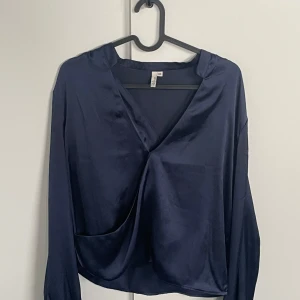 Mörkblå omlottopp från NLY Trend - Snygg mörkblå omlottopp från NLY Trend i ett glansigt material. Toppen har långa ärmar och en v-ringad design som ger en elegant look. Perfekt för att lyfta din outfit med en touch av stil.