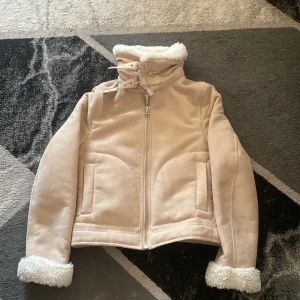 Beige jacka med fuskpälsfoder - Snygg beige jacka med mjukt fuskpälsfoder och dragkedja framtill. Jackan har långa ärmar och stiliga spännen vid kragen för extra detalj. Perfekt för kyligare dagar!