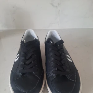 Svarta sneakers från Fred Perry - Snygga svarta sneakers från Fred Perry med vit sula och klassisk logga på sidan. Skorna har snörning och är tillverkade i skinn, vilket ger en stilren look. Fåtal ggr använda .
