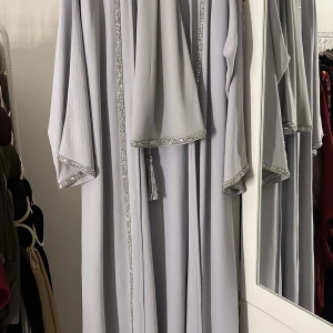Abaya  med glittriga detaljer - Elegant ljusgrå abaya med glittriga kanter och ett knytband i midjan. Klänningen har långa ärmar och en avslappnad passform. Passande sjal och underklänning följer med. Användes endast en gång! (Kan fraktas) 