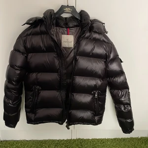 Moncler Maya - Säljer en snygg svart dunjacka från Moncler. Jackan har en quiltad design med dragkedja och huva. Perfekt för kalla dagar med sin varma och stilrena look. Den har långa ärmar och praktiska fickor med dragkedjor.