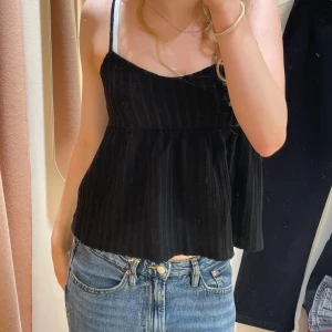 Svart ribbad linnetopp - Säljer en svart ribbad linnetopp med smala axelband och knytning i ryggen. Toppen har en luftig passform och är perfekt för varma dagar. Den är stilren och lätt att matcha med olika outfits.