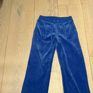 Blå mjukisbyxor från Juicy Couture - Snygga blå mjukisbyxor från Juicy Couture i en mjuk och glansig finish. Byxorna har en dragsko i midjan och broderad logga på fickan. Perfekta för en avslappnad stil.
