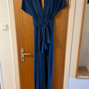 Mörkblå långklänning med knytband - Elegant mörkblå långklänning med v-ringning och knytband i midjan. Klänningen har kortärmade ärmar och ett flytande fall som ger en vacker siluett. Perfekt för en stilfull kväll.