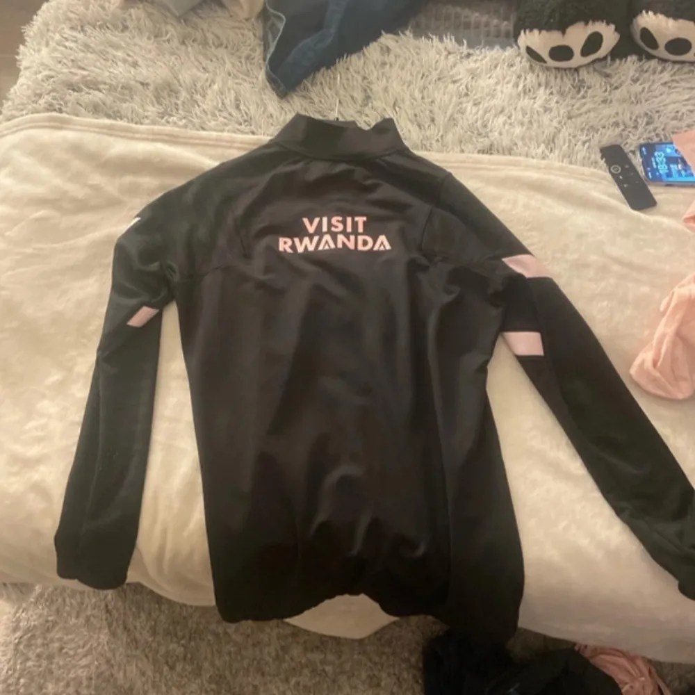 Snygg svart långärmad fotbollströja från Nike med rosa detaljer och PSG-logga. Tröjan har en halv dragkedja framtill och texten 'Accor Live Limitless' samt 'Visit Rwanda' på baksidan. Perfekt för fotbollsfans!. Neuletakit & Villapaidat.