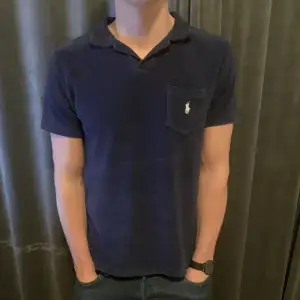 Snygg mörkblå pikétröja från Ralph Lauren med klassisk krage och korta ärmar. Tröjan har en broderad logga på bröstfickan. Perfekt för en stilren look.