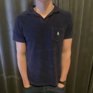 Mörkblå pikétröja från Ralph Lauren - Snygg mörkblå pikétröja från Ralph Lauren med klassisk krage och korta ärmar. Tröjan har en broderad logga på bröstfickan. Perfekt för en stilren look.