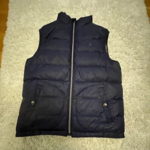 Dunväst i blå och grå - Säljer två snygga dunvästar i blått och beige. Båda har dragkedja framtill och praktiska fickor med knappar. Perfekta för kyligare dagar och lätta att matcha med olika outfits.