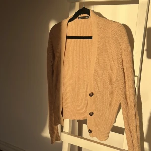Beige stickad kofta med knappar - Säljer en mysig beige stickad kofta med stora svarta knappar. Ger en stilren look. Koftan har långa ärmar och en klassisk passform.
