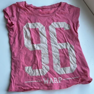 Rosa t-shirt med tryck - Säljer en rosa t-shirt med stort vitt tryck av siffran 96 på framsidan. T-shirten är kortärmad och har en rund halsringning. Perfekt för en avslappnad stil. VET EJ VAD DET ÄR FÖR STORLEK MEN FÖR MINDRE BARN