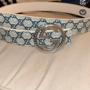GG gucci bälte mönstrat med silver badge  - Snyggt vitt bälte från Gucci med ett marinblått mönster på. Bältet har en stor, rund spänne i silver med det ikoniska GG-emblemet. Perfekt för att ge en lyxig touch till din outfit.