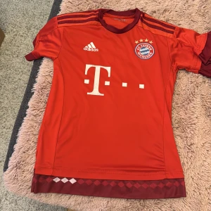 Röd Bayern München fotbollströja - Säljer en röd Bayern München fotbollströja från Adidas med korta ärmar. Tröjan har klubbens emblem och sponsorlogga på framsidan samt spelarnamnet Götze och nummer 19 på baksidan. Perfekt för fans av laget!