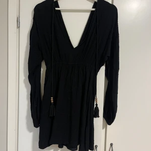 Svart klänning med v-ringning - Elegant svart klänning med djup v-ringning och långa ärmar. Klänningen har dekorativa snören med tofsar framtill och en smickrande passform. Perfekt för en stilren look.