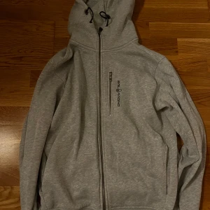 Grå hoodie från Sail Racing - Snygg grå hoodie från Sail Racing med dragkedja och justerbar huva. Perfekt för en avslappnad stil. Den har en praktisk bröstficka med dragkedja och logotyp på bröstet. I användt men fint skick. Storlek M