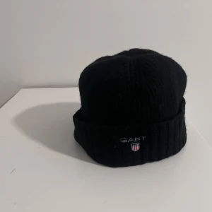 Svart ribbad mössa från Gant - Snygg svart ribbad mössa från Gant med broderad logotyp framtill. Perfekt för kyliga dagar med sin mjuka och varma insida i grått. Stilren design som passar till det mesta.