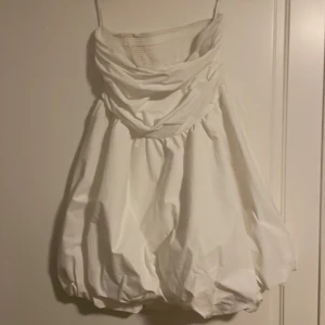 Vit studentklänning  - Säljer en unik vit studentklänning med axelbandslös design. Klänningen har en voluminös form och draperad överdel som ger en elegant look. Perfekt för student.