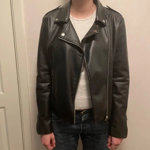 Svart skinnjacka med dragkedja - Snygg svart skinnjacka i fuskskinn med asymmetrisk dragkedja och klassisk bikerstil. Jackan har långa ärmar och stilrena detaljer som knappar vid kragen. (Obs - inte äkta läder)