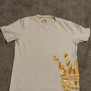 Vit t-shirt med gulddetalj från MR originals  - Snygg vit t-shirt från MR med en unik gulddetalj i form av en krona på sidan. Perfekt för dig som vill sticka ut med stil. T-shirten har korta ärmar och sitter som M perfekt om man är lite längre och har mellan L och M.