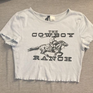 Ljusblå croppad t-shirt från H&M Divided - Säljer en ljusblå croppad t-shirt från H&M Divided med trycket 'The Cowboy Ranch' och en cowboy på häst. T-shirten har korta ärmar med vågig kant och är perfekt för en avslappnad stil.