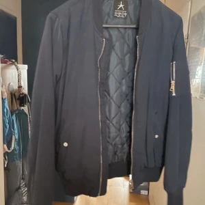 Blå bomberjacka med dragkedja - Snygg blå bomberjacka från Alpha Industries med dragkedja och quiltad insida. Jackan har en ficka med dragkedja på ärmen och ribbade muddar vid ärmslut och nederkant. Perfekt för en cool och avslappnad stil.