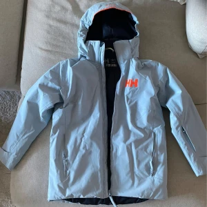 Ljusblå jacka från Helly Hansen - Säljer en ljusblå jacka från Helly Hansen med huva och dragkedja. Jackan har två fickor med dragkedjor och en orange HH-logga på bröstet. Perfekt för kyliga dagar. Passar någon som är runt 150 cm. 