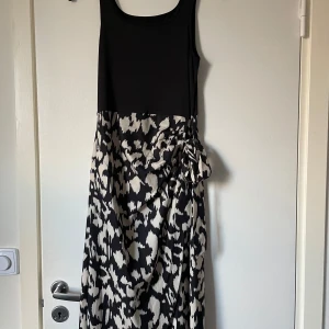 Svart och vit mönstrad klänning - Elegant klänning med svart överdel och mönstrad kjol i svart och vitt. Klänningen har en ärmlös design och en vacker drapering på sidan. Perfekt för en stilfull look.