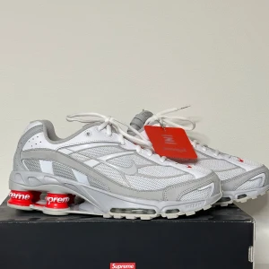 Nike x Supreme sneakers - Snygga Nike x Supreme sneakers i vitt och grått med röda detaljer. Skorna har en unik design med Supreme-loggan på hälen och en bekväm sula med röda dämpningspunkter. Perfekta för den stilmedvetna.