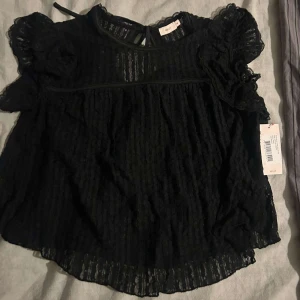 Svart blus från Nelly - Säljer en svart blus från Nelly med volangdetaljer och knytning i ryggen/nacken. Den är aldrig använd!! FRAKTEN STÅR MAN FÖR SJÄLV