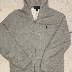 Grå hoodie från Polo Ralph Lauren - Säljer en grå hoodie från Polo Ralph Lauren med dragkedja och huva. Den har ett litet broderat märke på bröstet och är fodrad med mjukt material. Perfekt för kyliga dagar! Tröjan är i storlek 14-16. Den har även inga defekter. 