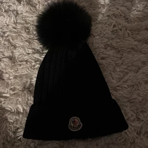 Svart mössa från Moncler - Snygg svart stickad mössa från Moncler med en stor pälsboll på.Aldrig andvänt