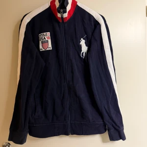 Mörkblå jacka från Polo Ralph Lauren - Säljer en mörkblå jacka från Polo Ralph Lauren med dragkedja och vita detaljer. Jackan har ett stort vitt Polo-logotyp på framsidan och USA-flagga med nummer 10 på baksidan. Tillverkad i bomull och polyester.
