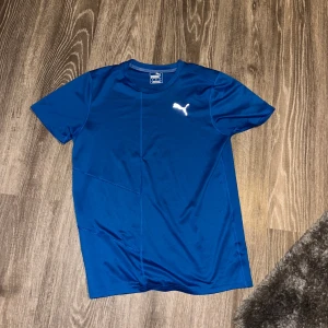 Blå t-shirt från Puma - Snygg blå t-shirt från Puma med korta ärmar och en vit logga på bröstet. Perfekt för träning eller vardagsbruk. Tillverkad i ett mjukt och bekvämt material.