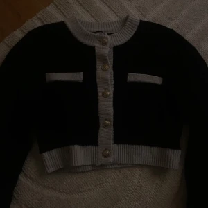 Svart och grå kofta från H&M - Säljer en snygg svart och grå kofta från H&M. Koftan har en croppad design med ribbade kanter och dekorativa knappar framtill. Perfekt för att lägga till en stilren touch till din outfit.