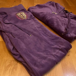 Lila hoodie och byxor från Juicy Couture - Säljer en lila hoodie och matchande byxor från Juicy Couture. Hoodien har dragkedja och snören vid huvan, medan byxorna har ett glittrigt emblem på sidan. Perfekt för en avslappnad stil.