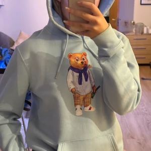 Ljusblå hoodie med björntryck - Säljer en ljusblå hoodie med ett coolt tryck av en björn i kläder. Hoodien har en klassisk passform med dragsko i huvan och en stor ficka framtill. Perfekt för en avslappnad stil!