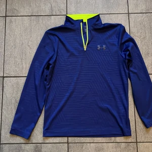 Blå tröja från Under Armour - Snygg blå tröja från Under Armour med dragkedja och limegrön insida vid kragen. Tillverkad i 100% polyester, perfekt för träning eller vardag. Långärmad design med diskret logga på bröstet.