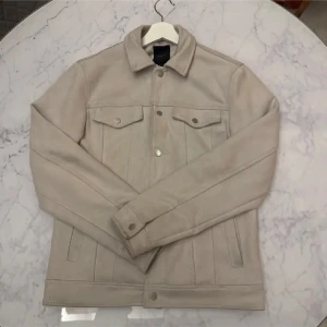 Beige jacka - Snygg beige jacka med klassisk krage och knappar framtill. Jackan har två bröstfickor med knappstängning och långa ärmar. Perfekt för en stilren look.