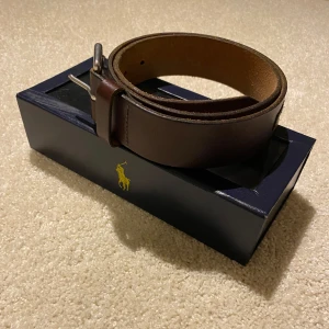Brunt bälte från Polo Ralph Lauren - Snyggt brunt bälte i skinn från Polo Ralph Lauren. Bältet har en stilren design med en silverfärgad spänne och märkets logga präglad på sidan. Perfekt för att ge en klassisk touch till din outfit.