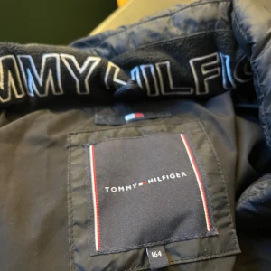 Svart pufferjacka från Tommy Hilfiger - Snygg svart pufferjacka från Tommy Hilfiger med dragkedja framtill. Jackan har en hög krage och är perfekt för kyligare väder. 